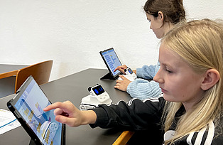 Zwei Kinder tippen auf Tablets und programmieren damit Roboter.