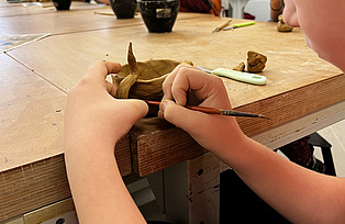 Ein Kind macht mit einem Werkzeug ein Muster in die Tonschale.