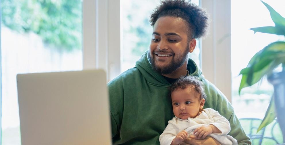 Young man holding a child while working with a laptop