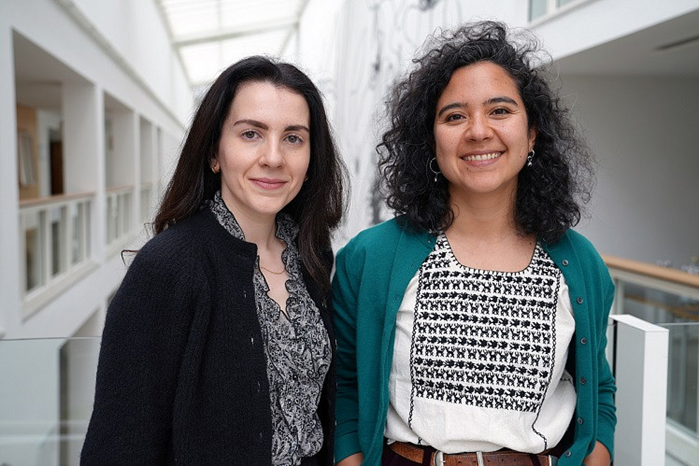 Dr. Pia Gutiérrez und Raquel Fernández Menéndez, zwei junge Frauen, stehen auf den Gängen der Universität und lächeln freundlich.