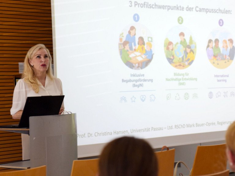 Blonde Frau steht hinter einem Rednerpult, im Hintergrund ist eine Powerpoint-Pr&auml;sentation zu sehen