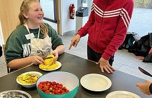 Auf einem Tisch steht eine Schale Erdbeeren und ein Kind schneidet eine Mango.