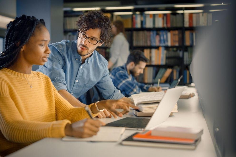 University Library: Gifted Black Girl uses Laptop, Smart Classma Dekoratives Bild, in dem eine Doktorandin an einem geöffneten Laptop sitzt und eine Frage an den Doktorand neben ihr sitzend bezüglich der Betreungsverereinbarung richtet.