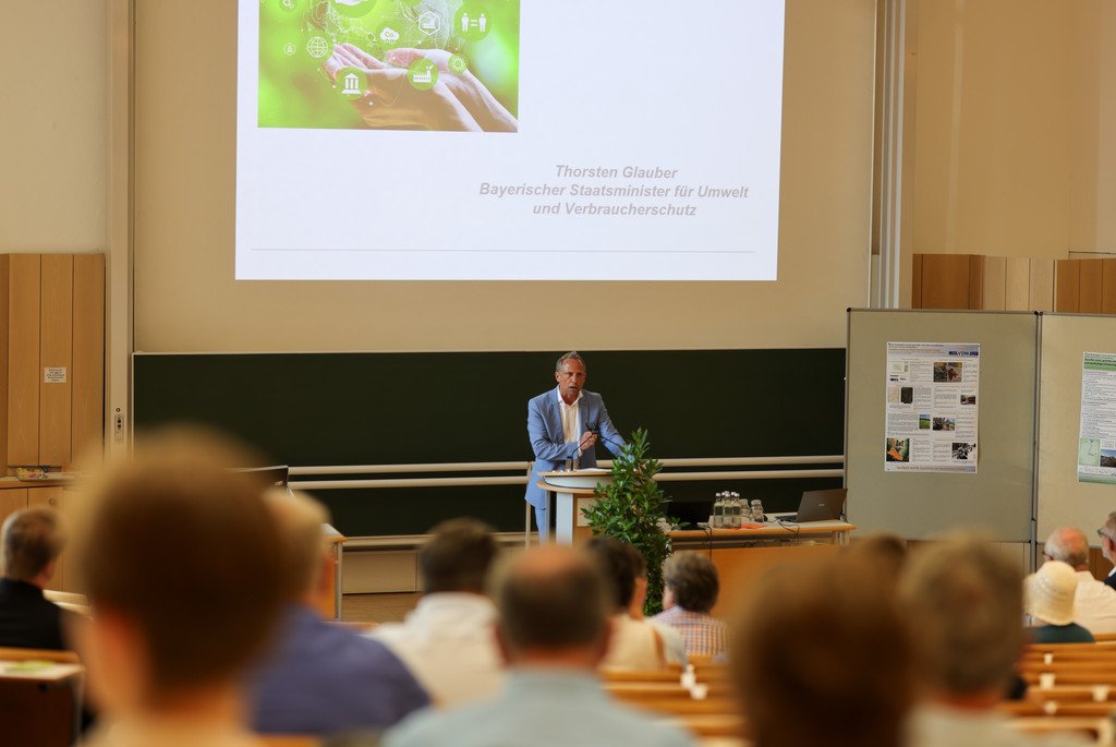 Staatsminister Thorsten Glauber steht an einem Rednerpult im Audimax der Universität Passau und spicht. Im Vorderung sieht man das sitzende Publikum mit dem Rücken zur Kamera.