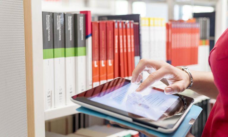 Studierende tippt auf Tablet, im Hintergrund im Bücher