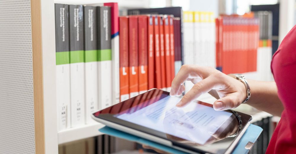 Studierende tippt auf Tablet, im Hintergrund im Bücher