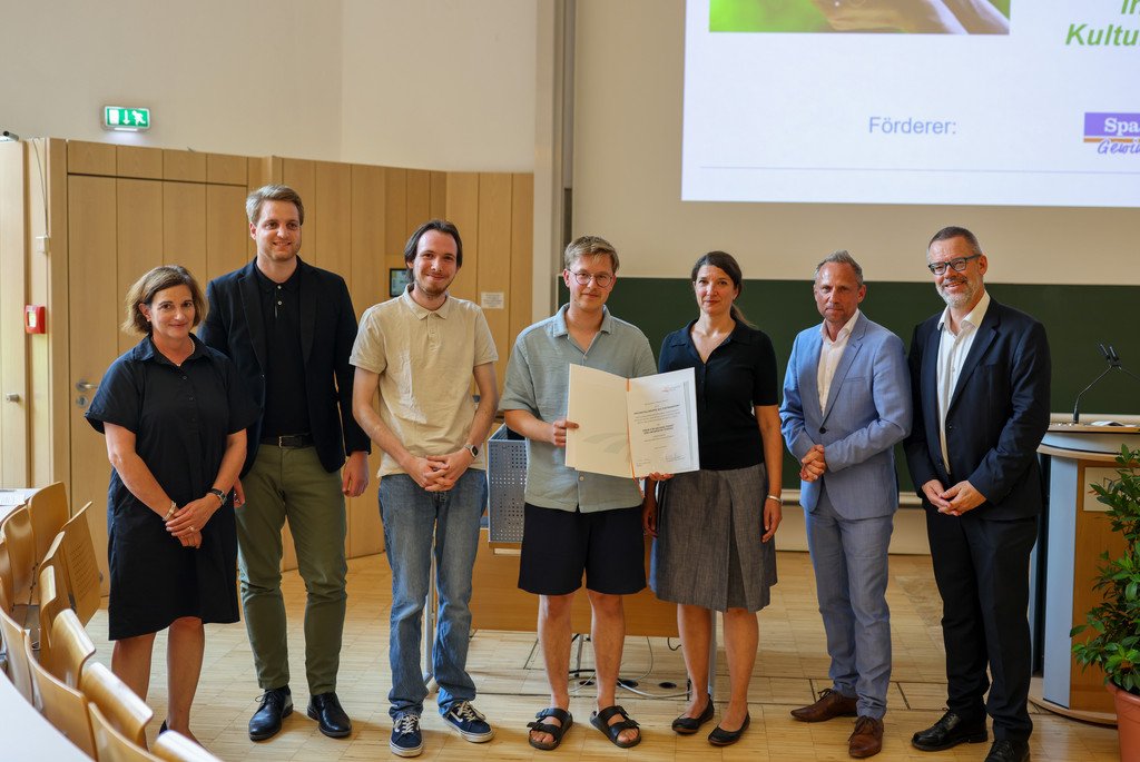 Der Kulturtransport an der Universit&auml;t Passau, vertreten durch Jonathan Halbauer, Clemens Diener und Dr. Stefanie Wehner erhalten am 3.7.2025 den Preis f&uuml;r Nachhaltigkeitsaktivit&auml;ten am Campus der Universit&auml;t Passau. Sie stehen zwischen Prof. Dr. Bettina Noltenius (Vizepr&auml;sidentin der Universit&auml;t Passau), Stephan Brandler (Leiter der Sparda-Bank-Filiale Passau), Staatsminister Thorsten Glauber (Bayerisches Staatsministerium f&uuml;r Umwelt und Verbraucherschutz) und Prof. Dr. Werner Gamerith (Vizepr&auml;sident der Universit&auml;t Passau). In der Mitte h&auml;lt Clemens Diener die Urkunde in die H&ouml;he.