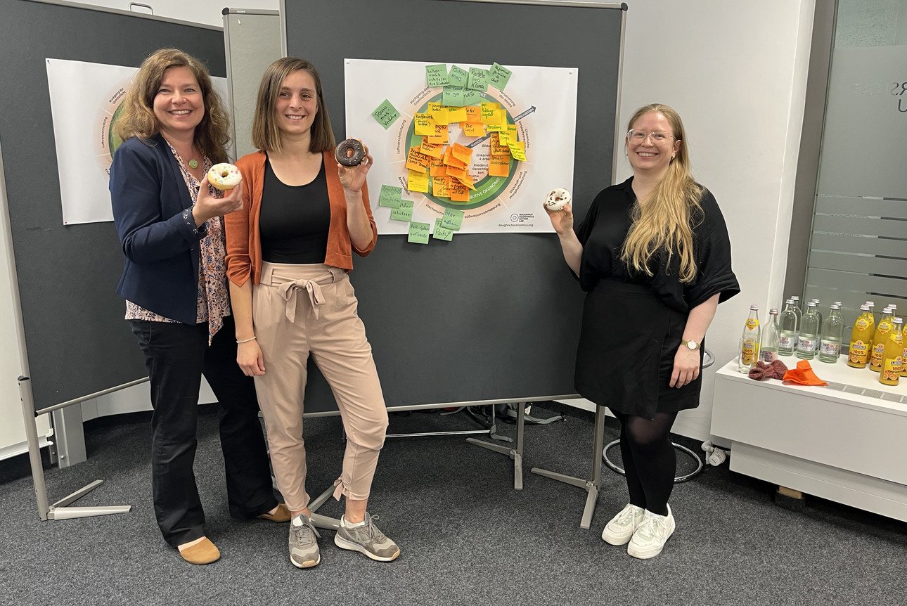Gruppenfoto mit Frau Dr. Annekatrin Mei&szlig;ner, Nelly Rahimy und Sina Kehrwieder, jeweils mit einem Donut in der Hand. Im Hintergrund eine Pinnwand mit Ideen zu &ouml;kologischen und sozialen Projekten f&uuml;r Passau.