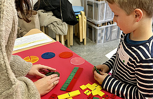 Mithilfe von unterschiedlich langen Holzplatten erklärt eine Workshopleitung einem Kind Brüche.