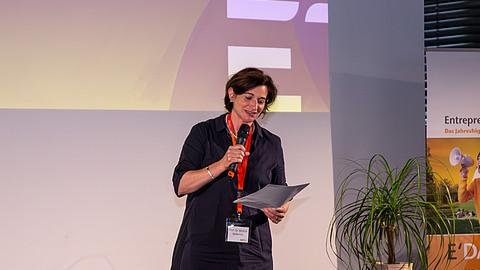 Prof. Dr. Carolin Häussler steht mit einem Mikrofon auf der Bühne und verkündet etwas von einem Zettel.