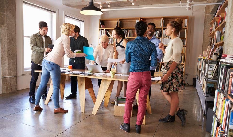 Several people standing around a table Eight people stand at a table and talk