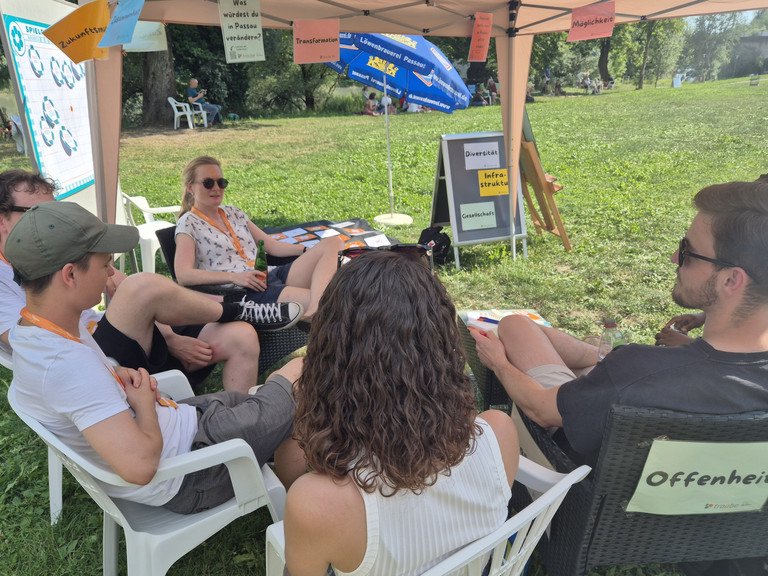 Gruppe von 5 Leuten sitzt auf Gartenst&uuml;hlen im Halbkreis und ist im Gespr&auml;ch