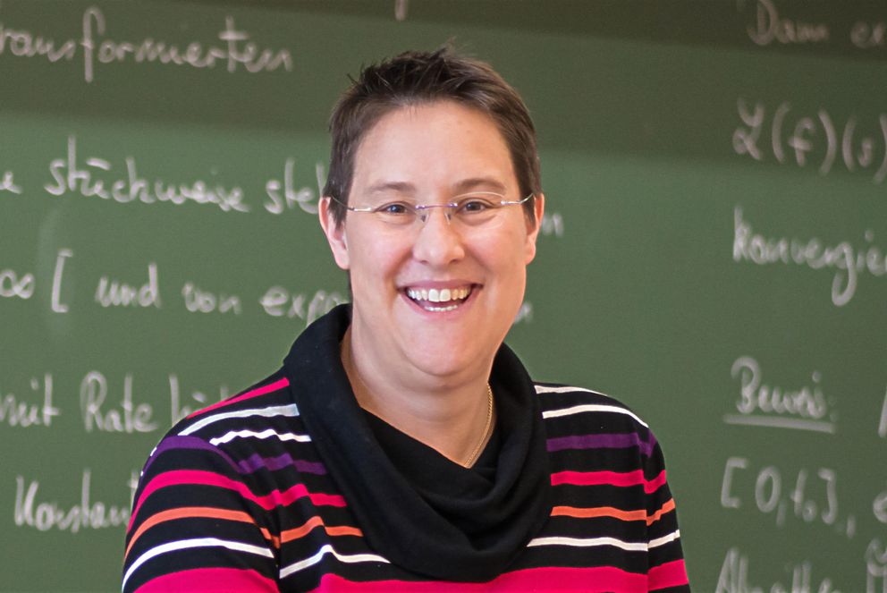 Prof. Dr. Brigitte Forster-Heinlein, Inhaberin der Professur für Angewandte Mathematik an der Universität Passau, steht herzlich lachend vor einer Tafel mit mathematischen Berechnungen. Sie hat sehr kurze, braune Haare und trägt ein lila-schwarzes Oberteil.