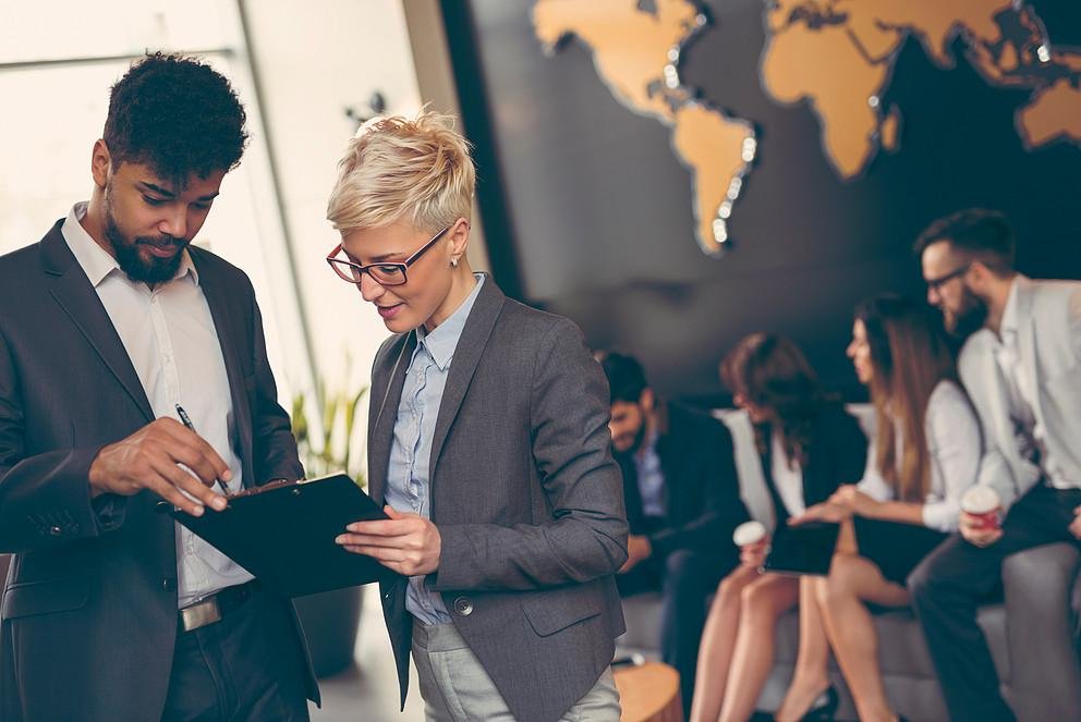 Business people working Eine internationale Gruppe junger Wissenschaftlerinnen und Wissenschaftler tauscht sich aus. Im Hintergrund sitzt eine Gruppe vor einer großen Weltkarte und unterhalt sich angeregt. Im Vordergrund hilft eine Mitarbeiterin des Welcome Centers einem internationalen Gastwissenschaftler bei einem Dokument.