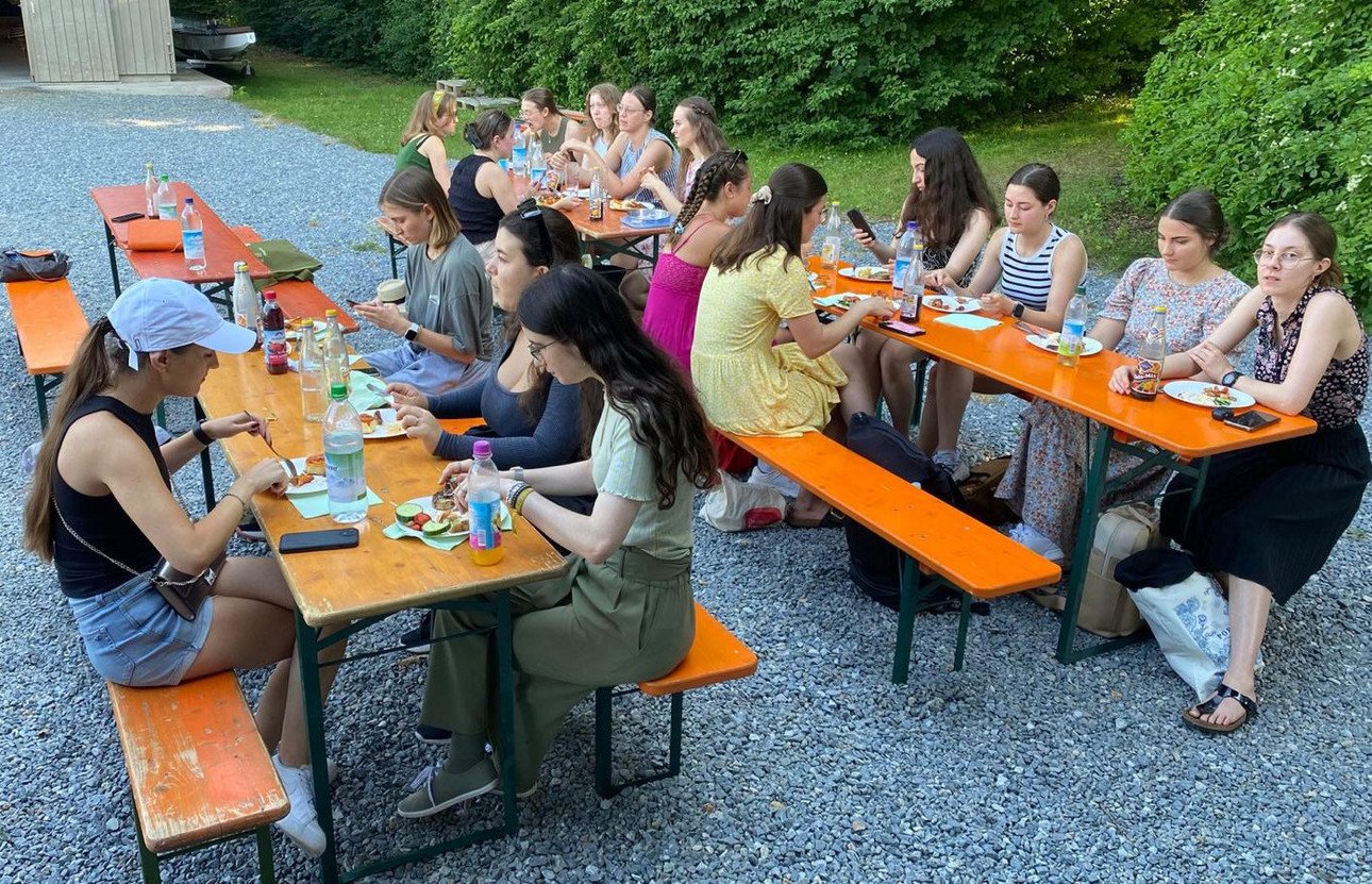 Stipendiatinnen und Stipendiaten sitzen bei Biertischen auf Bierb&auml;nken vor dem Bootshaus der Universit&auml;t Passau. Das Abschlussevent bei k&uuml;hlen Getr&auml;nken und gegrillten Speisen fand im Freien statt.
