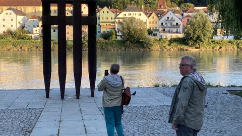 [Translate to Tschechisch:] Denkmalansicht mit Blickrichtung Inn bei abendlichem Licht aus ca. 10m Entfernung, mit zwei Besuchern davor.