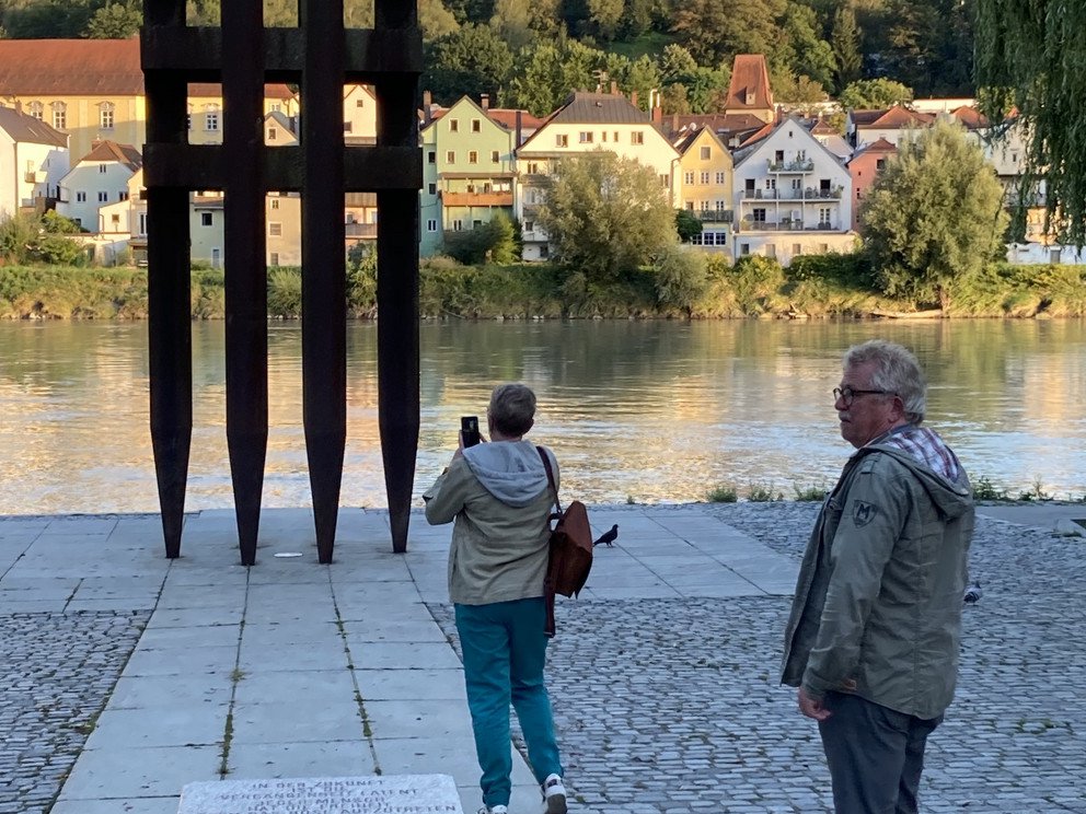 Denkmalansicht mit Blickrichtung Inn bei abendlichem Licht aus ca. 10m Entfernung, mit zwei Besuchern davor.