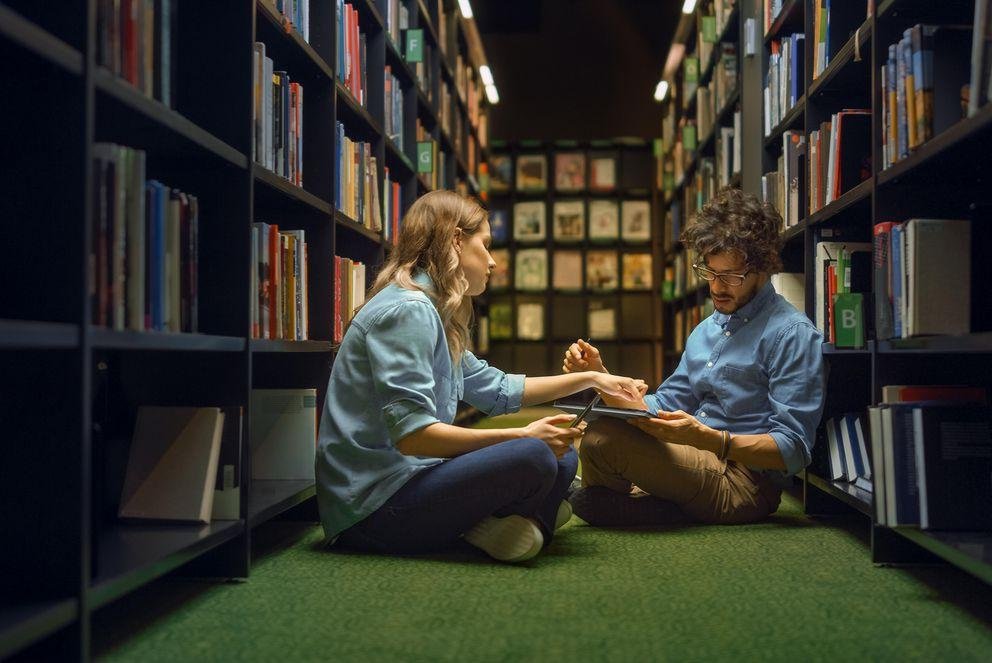 Two doctoral students are sitting on the green-carpeted floor of their library. They have both completed their dissertations and are thinking together about how best to publish them. The doctoral student is researching in his laptop. The other doctoral student reads along and gives him suggestions.