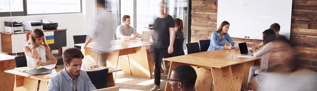 Großraumbüro mit mehreren Personen, die an einem Laptop sitzend arbeiten