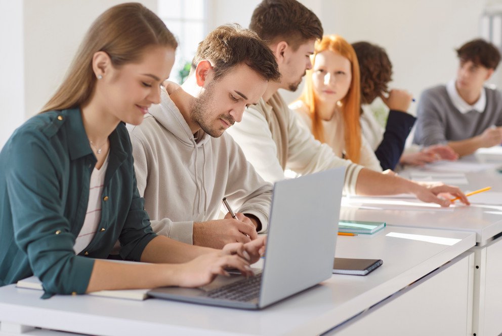 Group of young confident college or university students sitting at the same desk in the classroom during a lesson doing educational project on laptop. Education and knowledge concept. Foto: Studio Romantic – stock.adobe.com