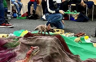 Kinder sitzen in einem Stuhlkreis und in der Mitte ist eine Landschaft mit Dinosauriern aufgebaut. 