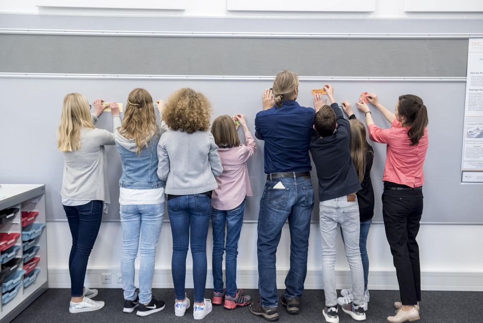 Schüler und Schülerinnen stehen mit einem Lehrer vor einer Wand, die mit Zetteln beklebt wird