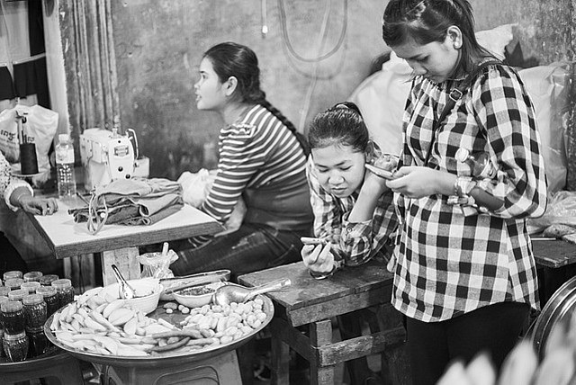 Schwarz-weiß Fotografie zeigt drei Mädchen mit Smartphones; Essen im Vordergrund, Nähmaschine im Hintergrund. Ort: Cambodia.