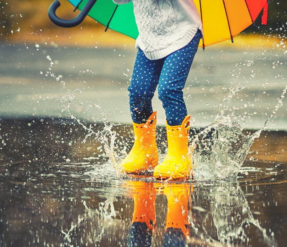 Füße eines Kindes in gelben Gummistiefeln, das im Regen über eine Pfütze springt