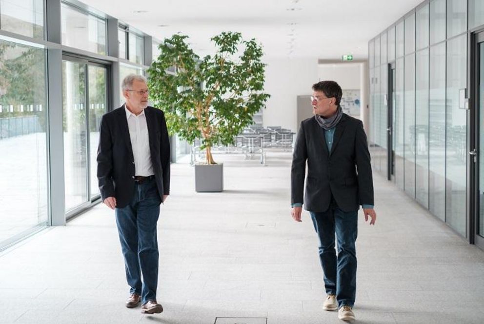 Der neue und der scheidende wissenschaftliche Leiter von TRIO Prof. Dr. Tomas Sauer und Prof. Dr. Burkhard Freitag laufen mit etwas Abstand nebeneinander her auf einem Gang der Universität.