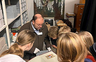 Die Kinder schauen zusammen mit dem Archivleiter ein Dokument an, auf  dem das bayerische Wappen zusehen ist an.