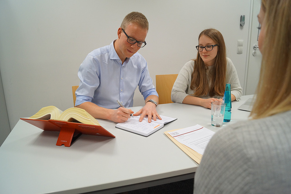 Darstellung einer Law Clinic Beratungssituation: Mann macht Notizen, vor sich ein Gesetzesbuch, daneben eine Kollegin.