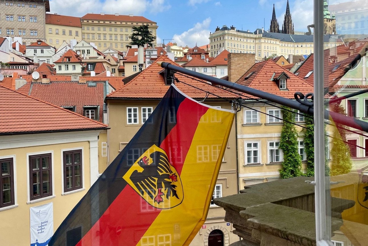 Auf diesem Bild sieht man eine deutsche Dienstflagge samt Adler, die aus der deutschen Botschaft in Prag nach drau&szlig;en weht.  Die Flagge ist an einem Metallmast befestigt, der schr&auml;g aus der Wand eines Geb&auml;udes ragt. Das Bild wurde aus einem Fenster des Geb&auml;udes aufgenommen. Im Hintergrund sieht man eine malerische Stra&szlig;e der Prager Altstadt, die mit Kopfsteinpflaster bedeckt ist. Links unten im Bild stehen zwei Menschen, die nach oben schauen und m&ouml;glicherweise ein Foto machen. In der N&auml;he sind mehrere Autos geparkt. Im Hintergrund sieht man eine Reihe von historischen Geb&auml;uden mit roten Ziegeld&auml;chern, die sich treppenartig den H&uuml;gel hinaufziehen. Einige dieser Geb&auml;ude sind hellgelb oder beige gestrichen. Am Horizont, hinter diesen Geb&auml;uden, ragen die spitzen T&uuml;rme der Prager Burg in den Himmel. Der Himmel ist klar und blau, mit einigen wei&szlig;en Wolken.