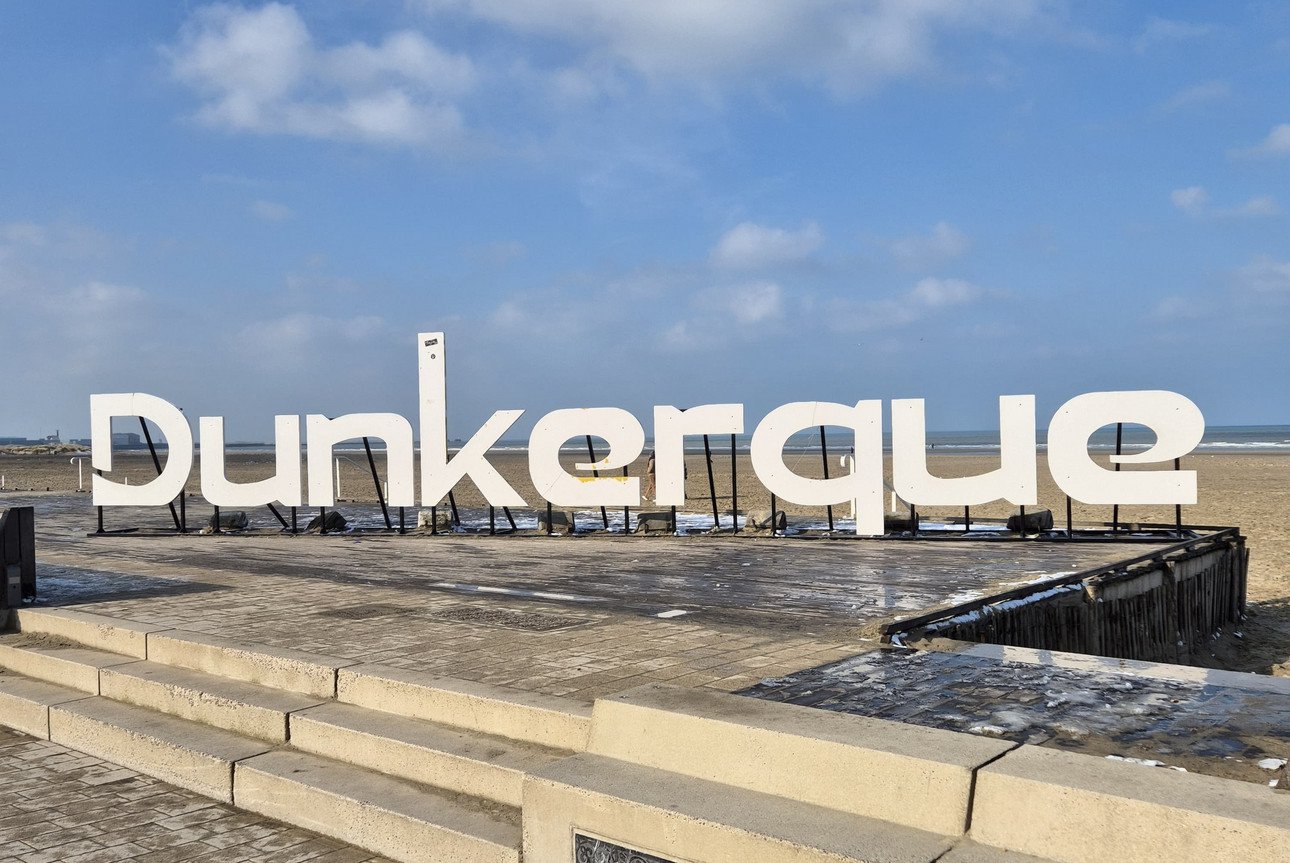 Gro&szlig;e "Dunkerque"-Schrift am Strand bei sonnigem Wetter mit Blick aufs Meer.