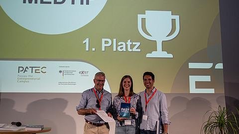 Ein Mann und zwei Studieren stehend lächelnd auf der Bühne und halten einen Preis in der Hand. 