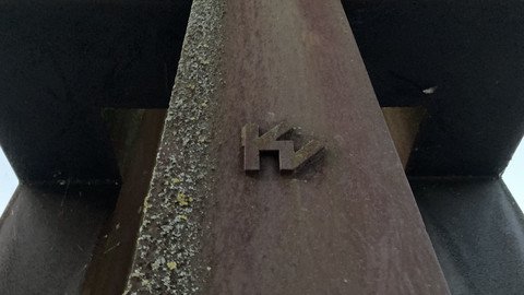 [Translate to Tschechisch:] Detailansicht der Skulptur, zu sehen ist ein angebrachtes plastisches nach außen stehendes Emblem, dass einer Verschmelzung eines Buchstaben "K" und eines um 120° gedrehten Buchstaben "M" oder eines um 45° gedrehten Buchstaben "W"
