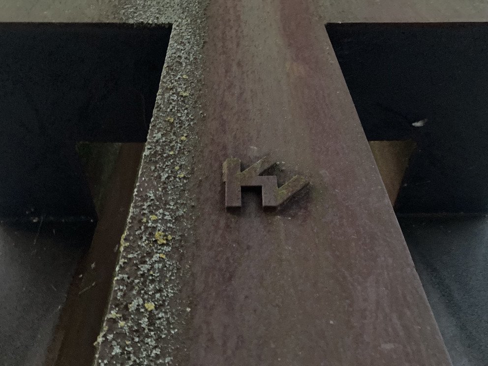 Detailansicht der Skulptur, zu sehen ist ein angebrachtes plastisches nach au&szlig;en stehendes Emblem, dass einer Verschmelzung eines Buchstaben "K" und eines um 120&deg; gedrehten Buchstaben "M" oder eines um 45&deg; gedrehten Buchstaben "W"