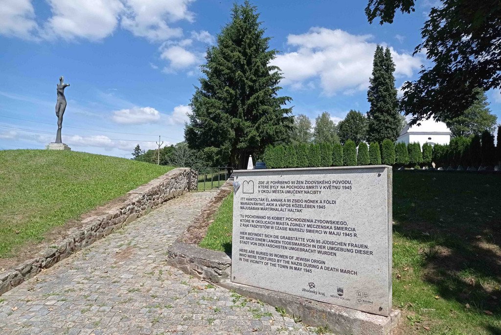 Ein Stein mit Aufschrift in mehreren Sprachen, im Hintergrund eine Statue. Aufgang zum Friedhof/K&aacute;men s n&aacute;pisy v několika jazyc&iacute;ch, v pozad&iacute; socha. Vchod na hřbitov.