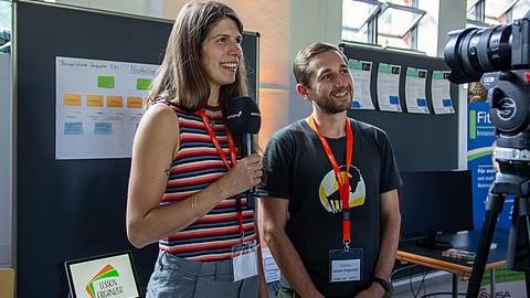 Das Team von Lesson Organizer steht mit einem Mikro vor ihrem Stand und lächelt in Richtung der Zuhörenden.