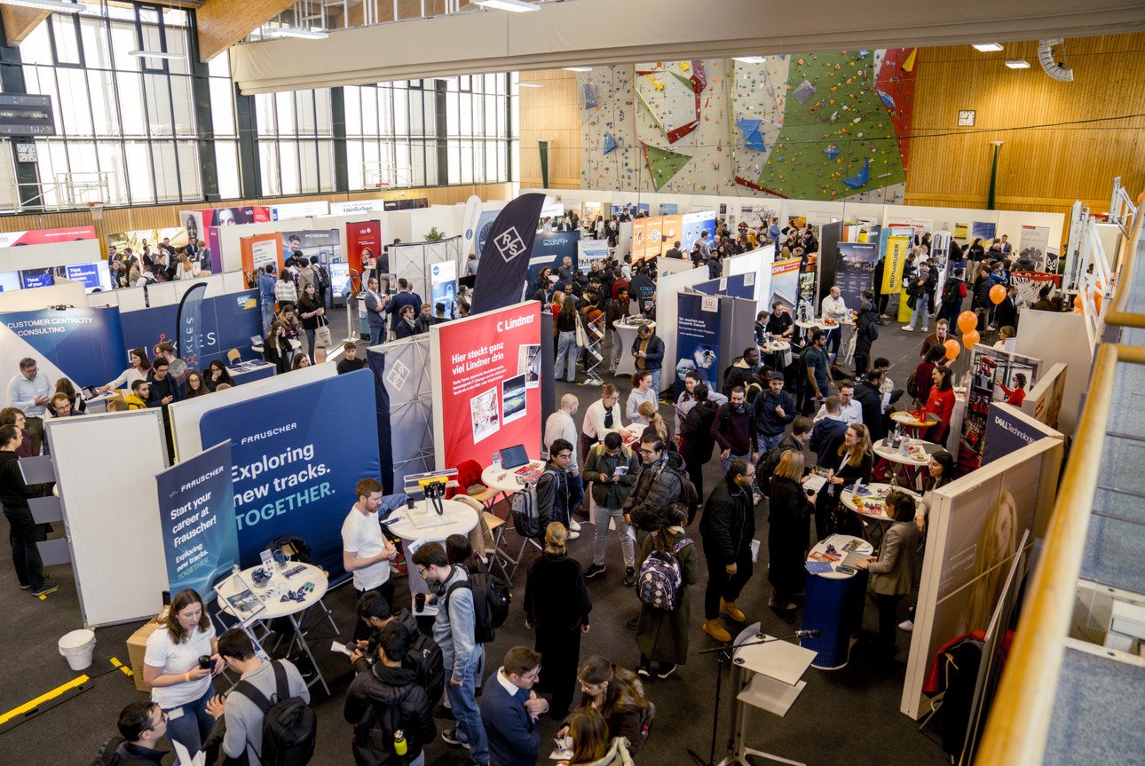 Auf dem Bild sieht man die Sporthalle der Universit&auml;t Passau aus einer erh&ouml;hten Perspektive. Die Halle ist voller Menschen und St&auml;nde der einzelnen Unternehmen, die bei der Campus meets Company 2024 ausgestellt haben.