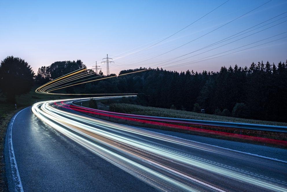 Eine Landstraße in der Dämmerung, auf der viele weiße und rote Lichter vorbeifahrender Autos zu Linien verschwimmen.