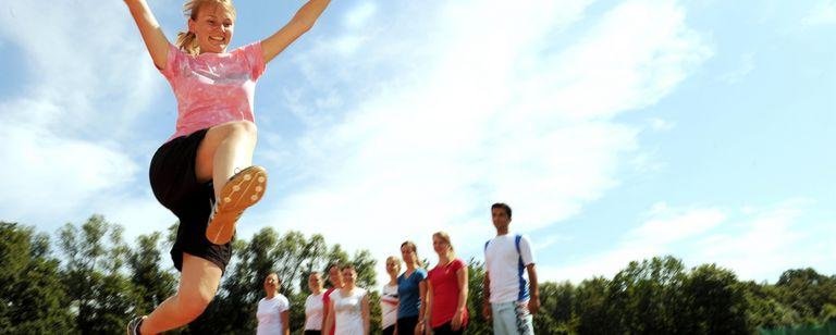 Das Bild zeigt eine Gruppe von Studierenden auf dem Sportplatz. Eine Studentin setzt zum Weitsprung an.