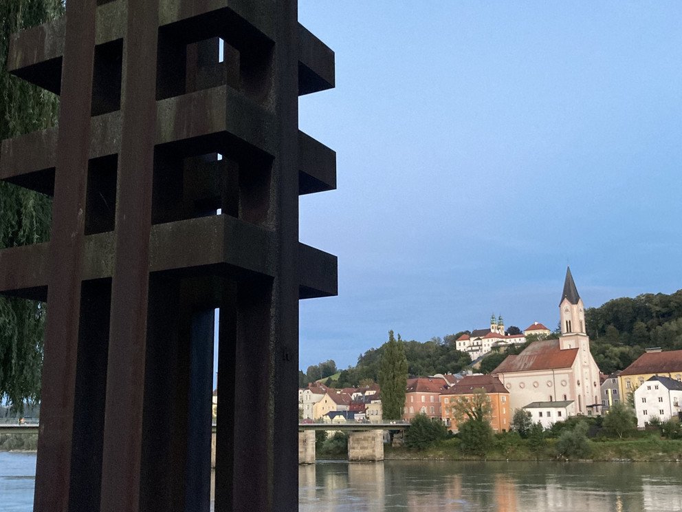 Ansicht der Skulptur aus etwa 5m Entfernung mit Blickrichtung Inn, im Hintergrund zu sehen die Kirche St. Gertraud