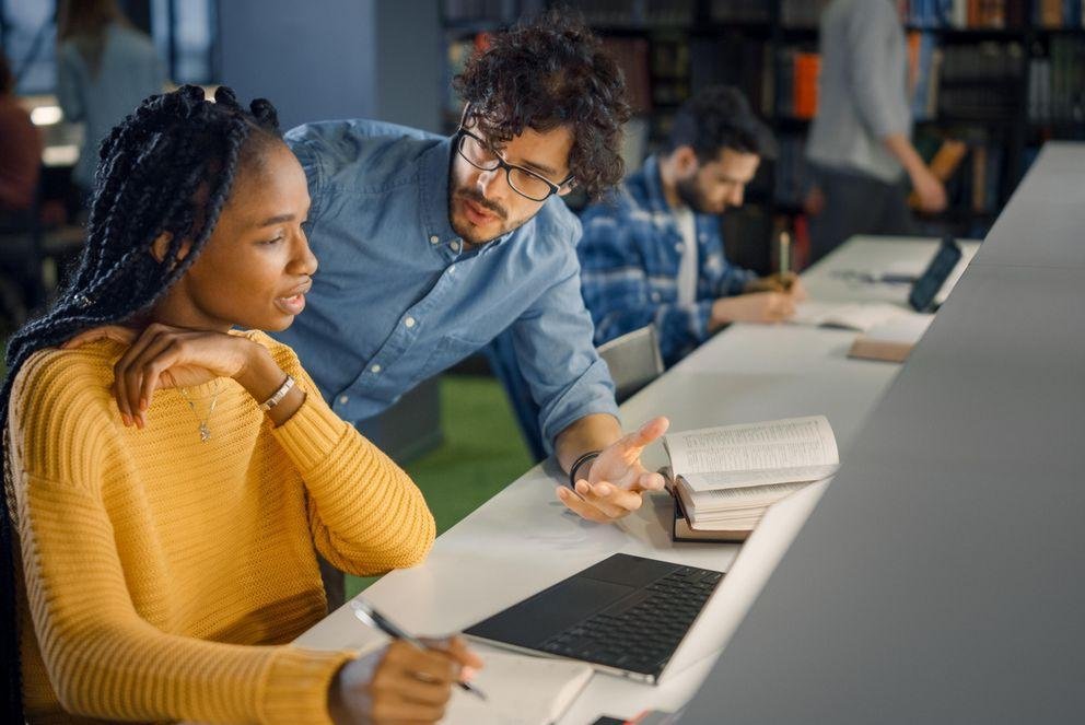 Decorative picture to break up the text on the German Act on Temporary Scientific Contracts (Wissenschaftszeitvertragsgesetz), showing a doctoral student sitting brooding over her laptop. Next to her sits another doctoral student who is explaining the German Act on Temporary Scientific Contracts to her.