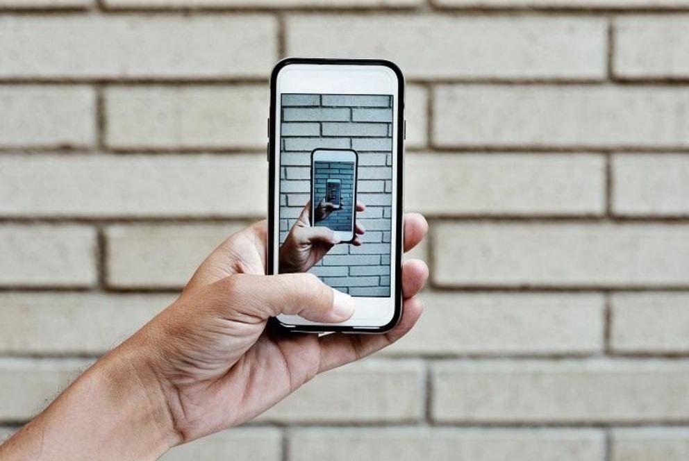 Ein Mensch hält ein Smartphone vor einer Backsteinmauer. Auf dem Display erscheint das selbe Bild, das wir gerade betrachten erneut. Darin erneut und so fort. 