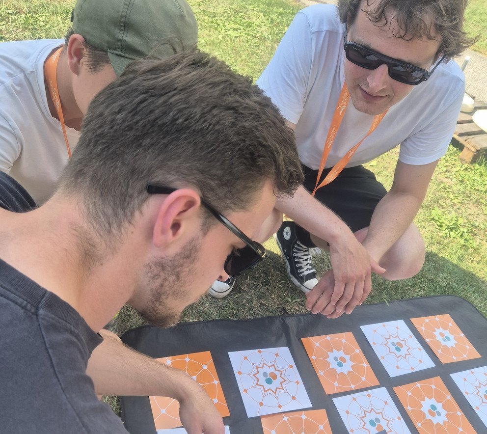 3 junge M&auml;nner sitzen auf dem Rasen am Boden und sind &uuml;ber orangene und wei&szlig;e Memory-Karten gebeugt und unterhalten sich