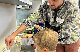 Eine Workshopleitung hilft einem Kind Milch abzumessen.