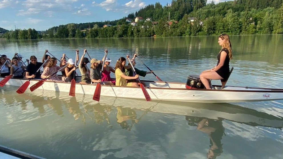 Stipendiatinnen und Stipendiaten rudern gemeinsam am Drachenboot am Inn. Eine Stipendiatin sitzt vorne an der Trommel. Im Hintergrund sieht man viele grüne Bäume.