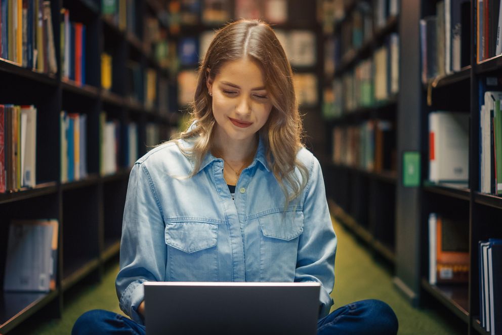 A doctoral student sits in the library surrounded by bookshelves. She is researching funding options for her doctorate. She is wearing a jeans shirt and medium-length brown hair.