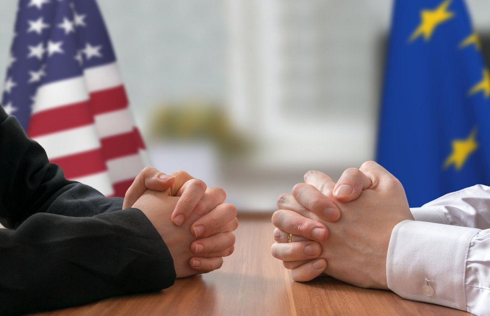 Symbolbild: Nahaufnahme von zwei Paar H&auml;nden mit amerikanischer und Europa-Flagge im Hintergrund