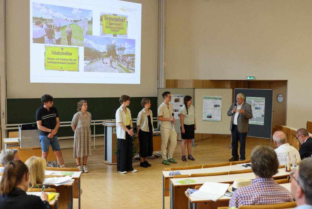 Prof. Dr. Hubert Weiger steht im Audimax der Universität Passau mit einem Mikrofon und spricht mit sechs Studierenden, die am Seminar "Grenzen überwinden" teilgenommen hatten.